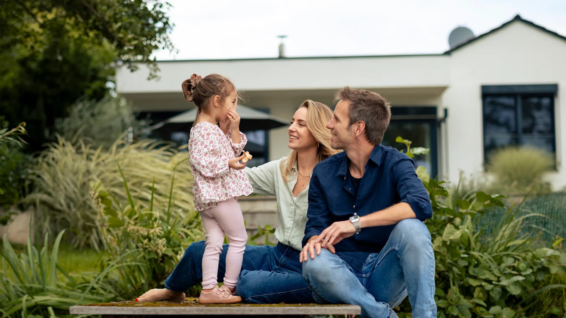 Familie mit Mutter, Vater und Tochter sitzen vor einem Haus 